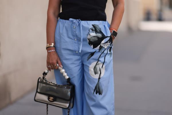 Woman in Vienna, wearing a black top from Zara, blue floral print pajama pants from Prada, various bracelets and rings, an Apple watch, and holding a transparent Chanel bag.